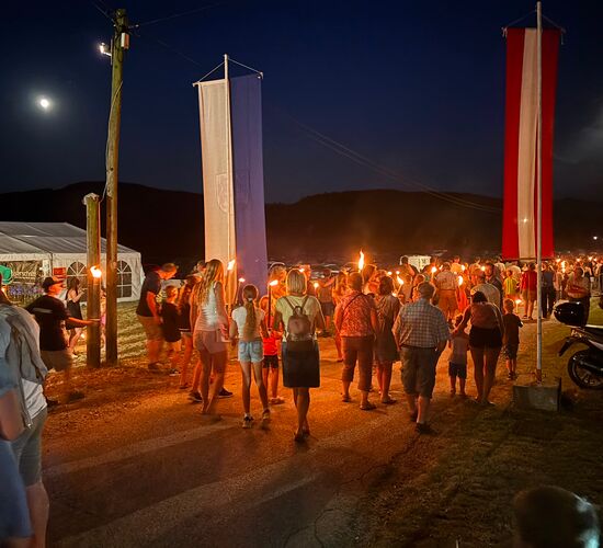 Eine Menschenmenge mit Fackeln feiert bei Nacht unter dem Mond, umgeben von Fahnen und festlicher Atmosphäre.